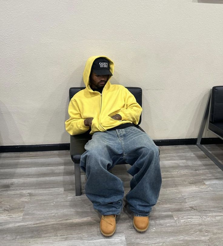 Persona de piel negra vistiendo sudadera amarilla oversize con pantalones jeans baggy, gorra negra y sentado en una silla